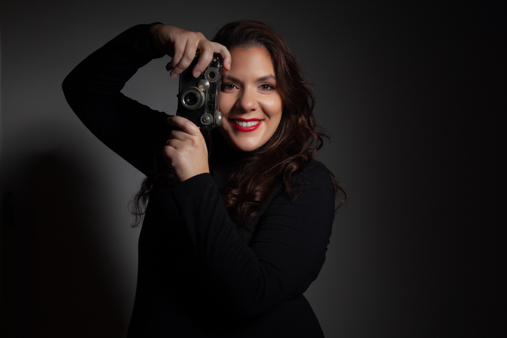 woman holding a vintage camera wearing a black turtleneck and smiling with red lipstick