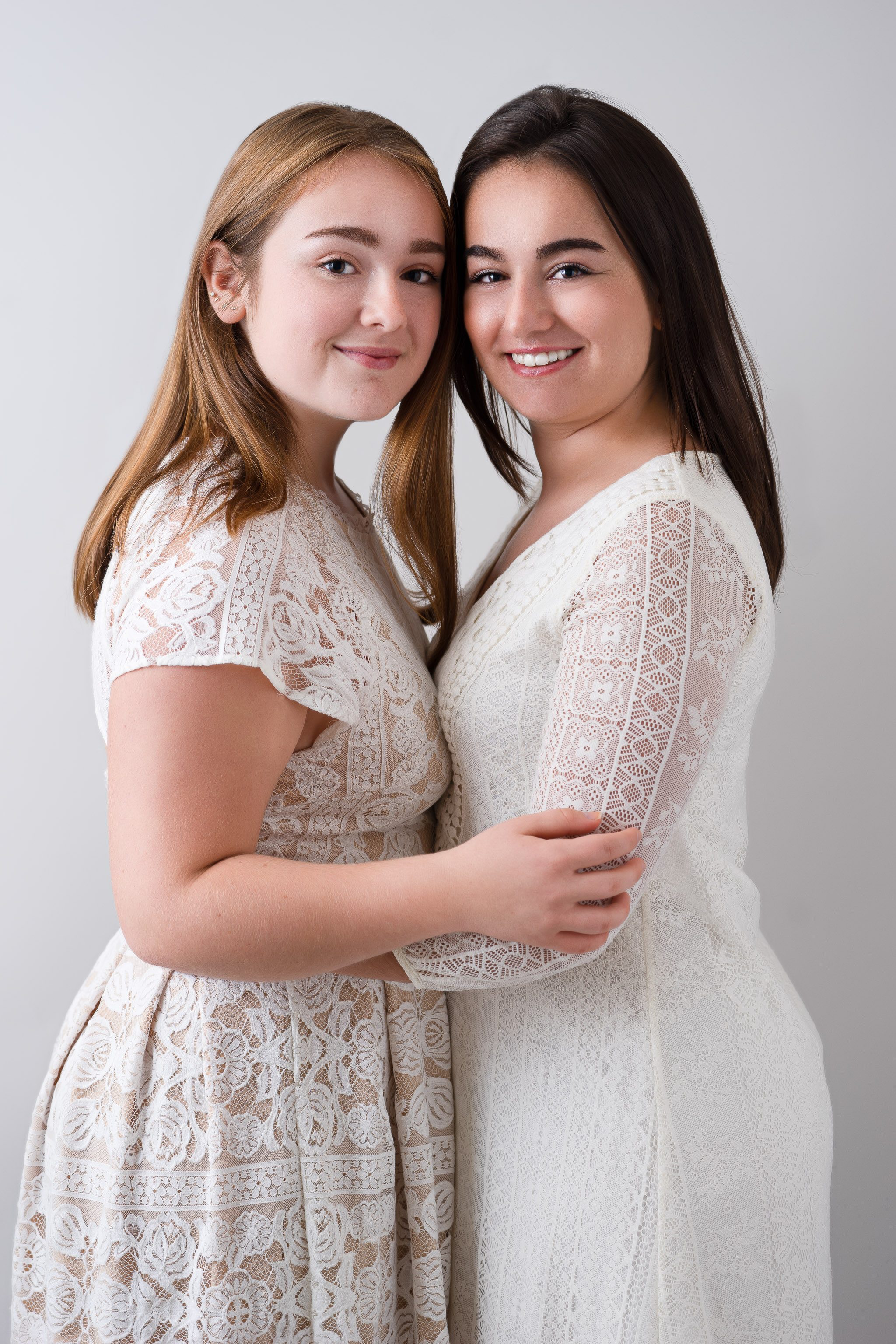 two teen sisters wearing white dresses in bright and airy lighting with arms around each other smiling joyfully at the camera