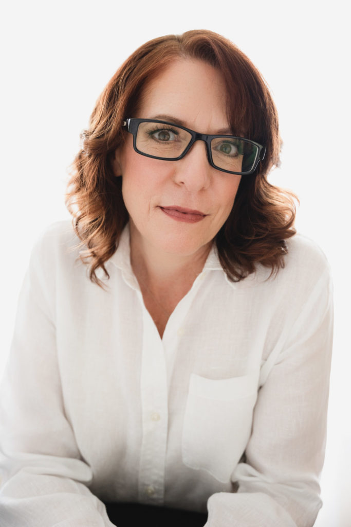 studious headshot of woman in a white button down shirt with medium length red hair and a half smile on her face but still serious. there is an interesting look in her eyes. She is happy but also serious
