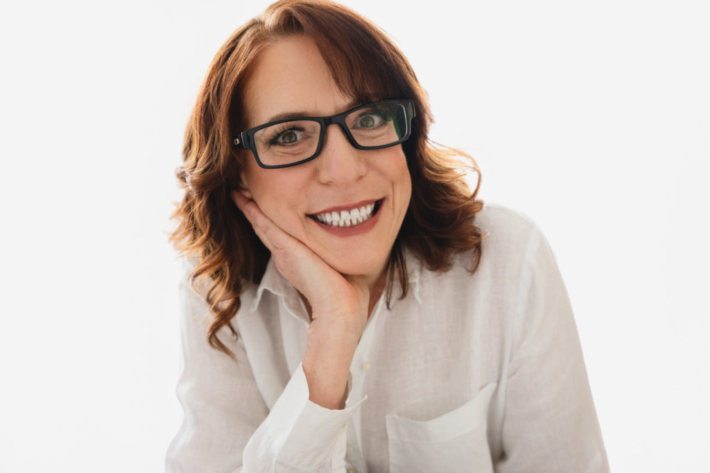 woman with longer red hair wearing black frame glasses and a white button down shirt posing for a studio headshot. Very happy with white teeth and a great smile. Hand on the chin pose