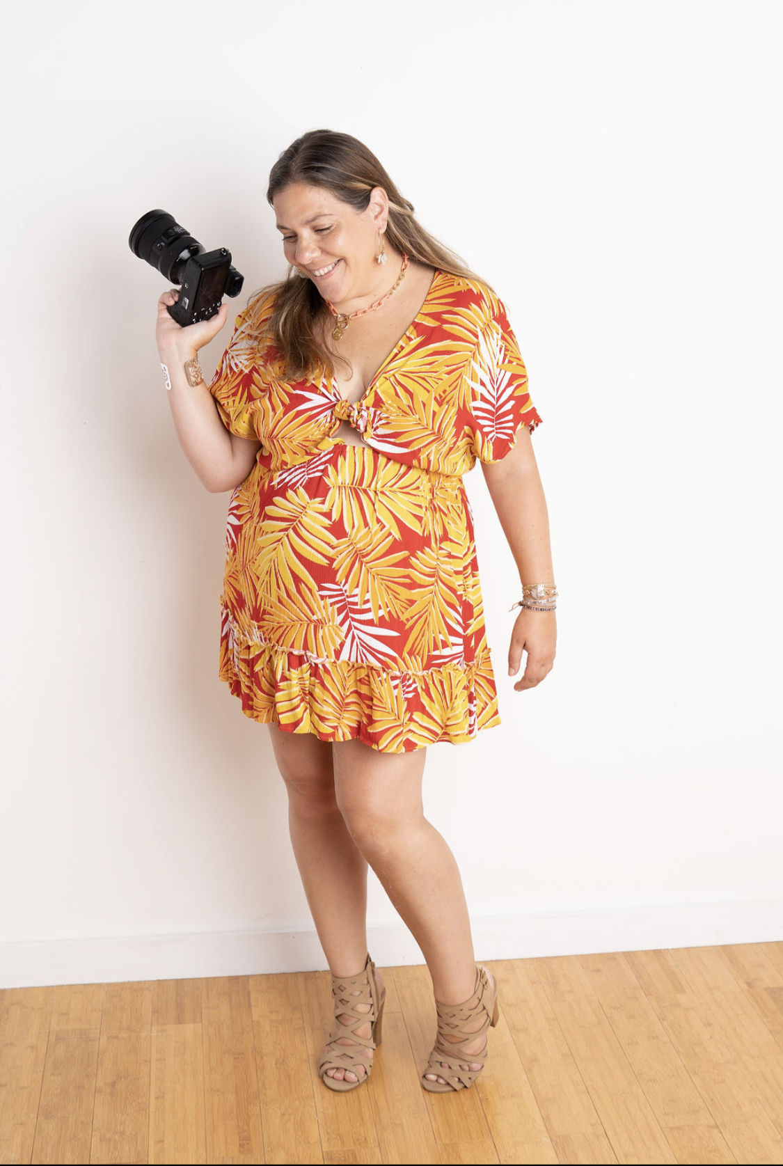 woman with long brown hair smiling as she looks down. Hawaiian yellow dress with red flowers. High heel beige sandles