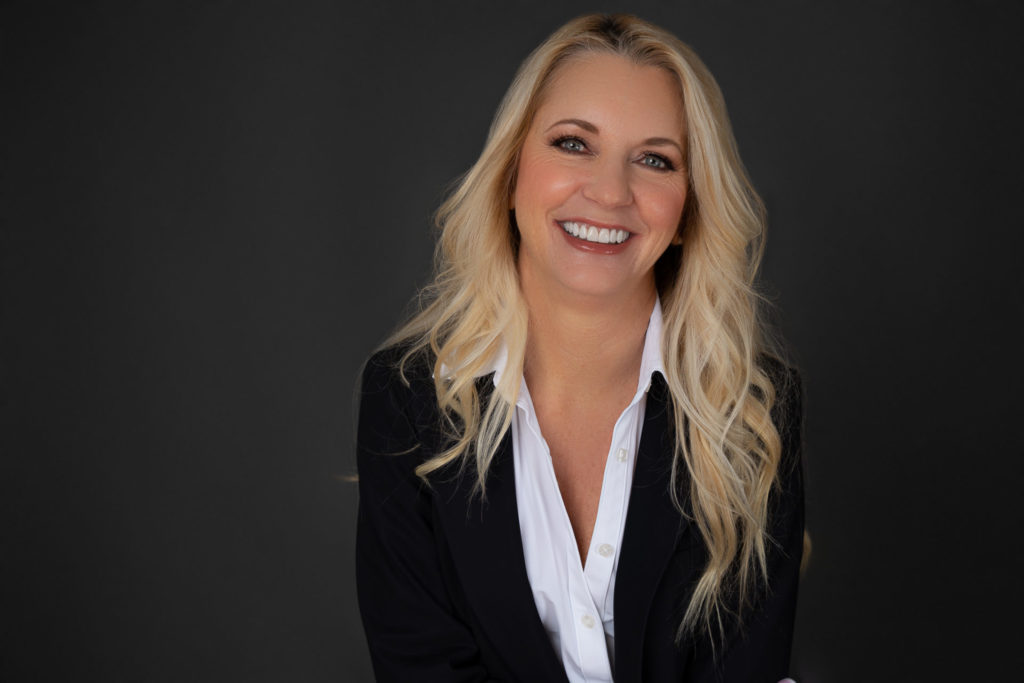 very pretty woman in her sixties with long blond hair wearing a business white colored shirt with black blazer, headshot, smiling woman, white teeth, blue eyes