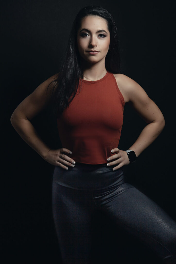 young woman with long black hair standing in a very strong warrior like pose wearing exercise clothing and has muscles and very active looking