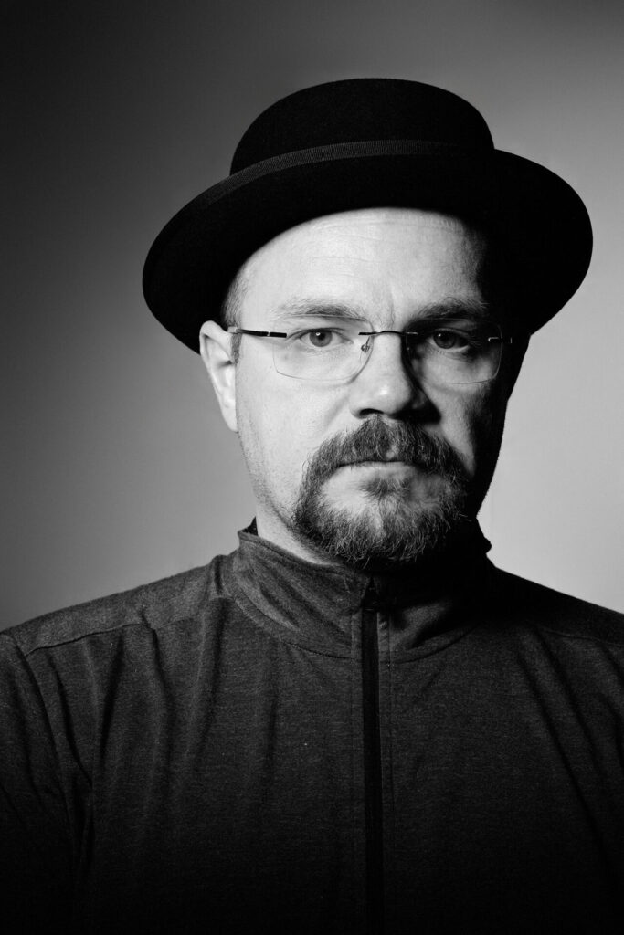 serious man with facial hair wearing glasses, port pie hat, black and white headshot middle age man