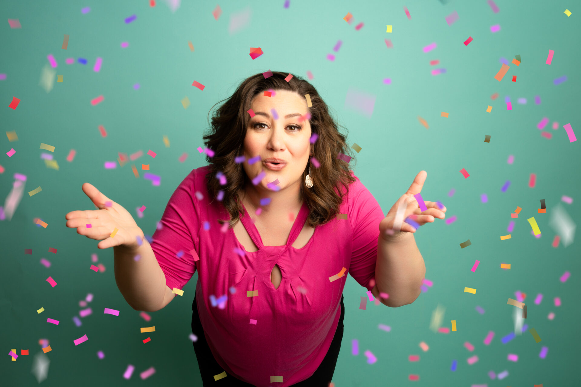 happy woman with short brown curly hair wearing a pink shirt blowing a kiss to the camera with confetti flying around