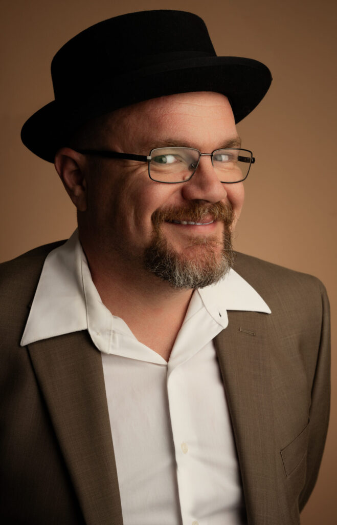 man in black hat smiling in suit, headshot, brown sports coat, white button down shirt, man with beard smiling with glasses