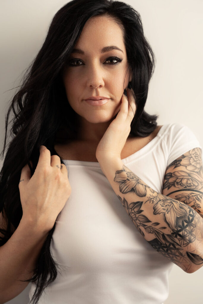 beautiful young women with long black hair wearing a simple white teeshirt has a tattoo sleeve and is staring boldly at the camera