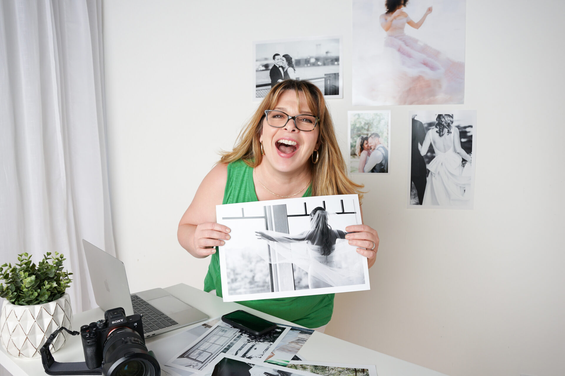 Behind-the-scenes photo of a photographer laughing in her Garden City studio while holding printed portraits, with camera and laptop on the desk