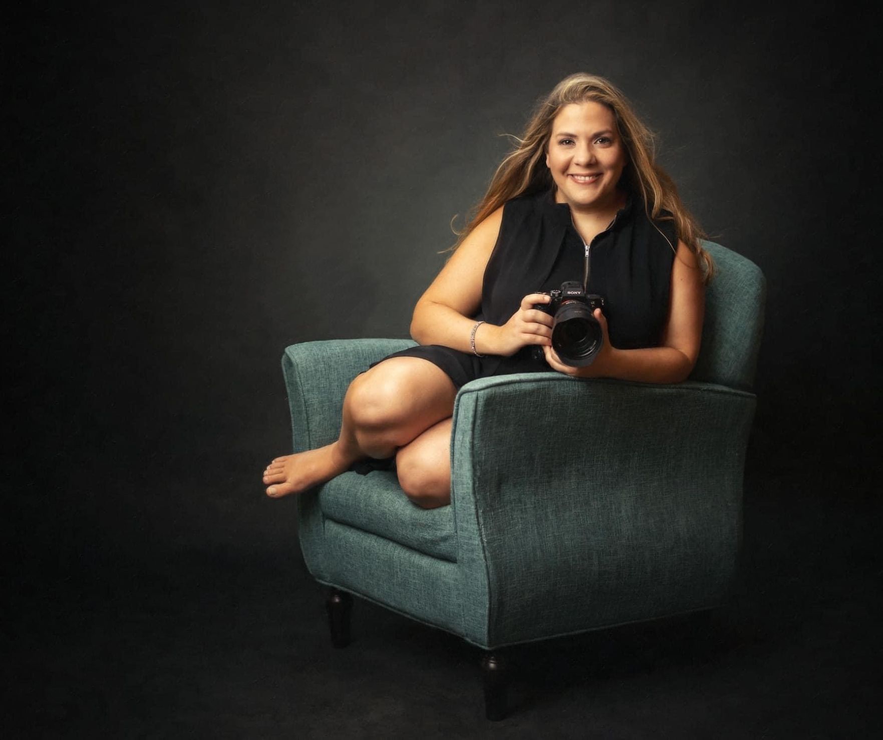 Portrait of Mary BeVard, professional portrait and personal branding photographer at The BeVard Studio in Farmingdale, NY, sitting on a teal chair holding a camera against a dark studio backdrop.