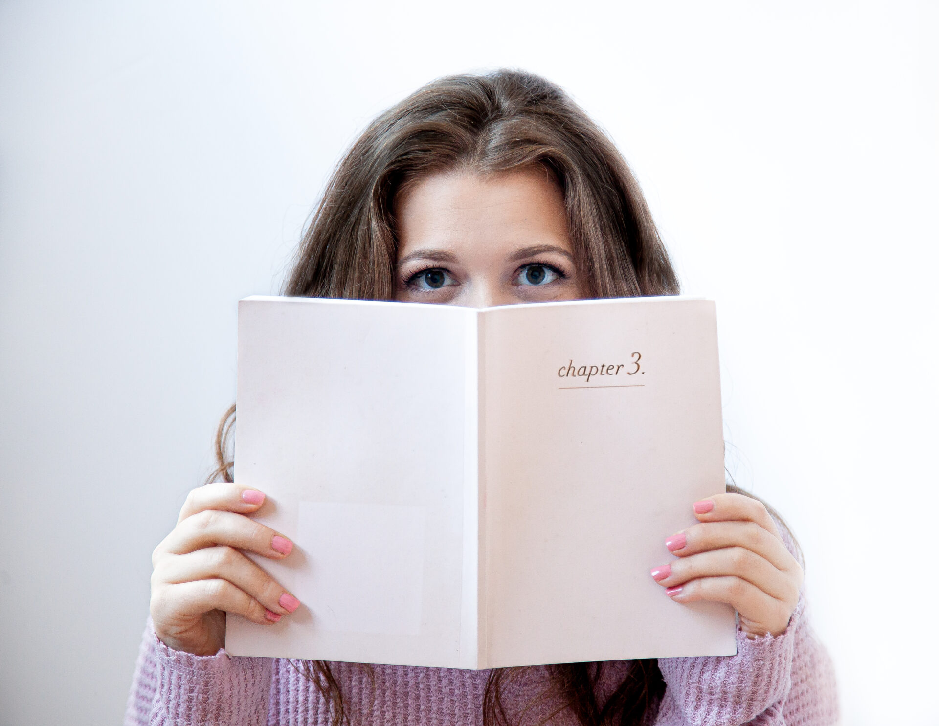 Personal branding portrait of a woman in a pink sweater holding a book titled “chapter 3,” photographed in a modern Long Island studio setting