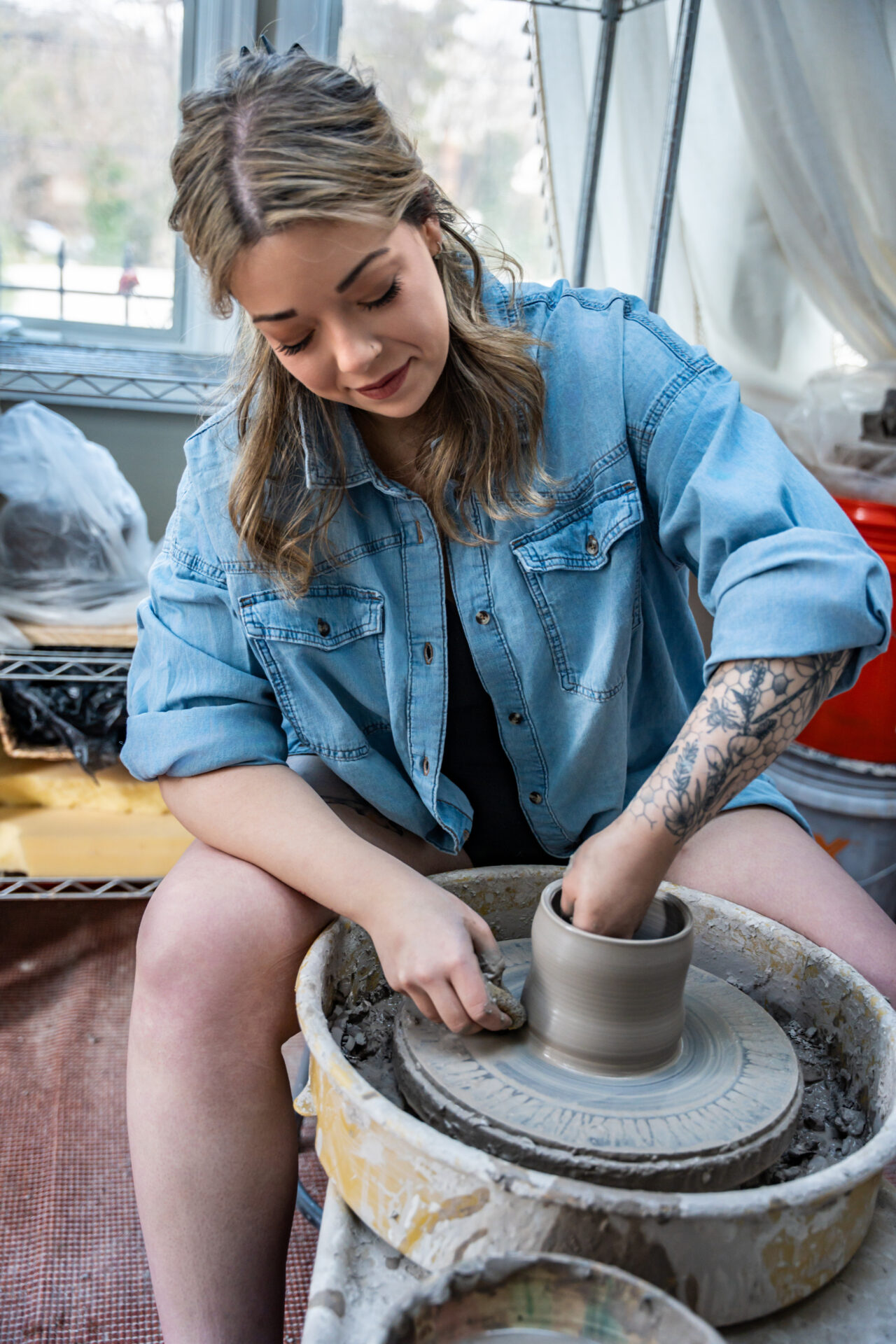 A female pottery artist with tattoos wearing a denim shirt, focused on shaping clay on a pottery wheel. Authentic lifestyle branding photography for creatives in Huntington, NY